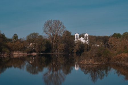 Old white stone catholic church reflected in calm water with blue sky looming over the dark trees. Historical gothic buildingの写真素材
