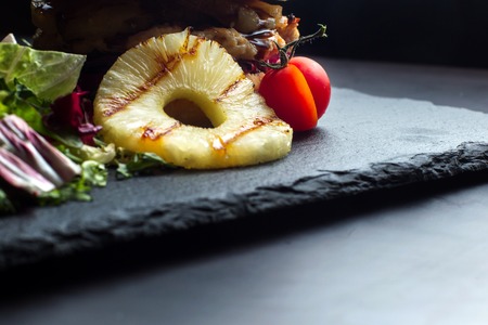 Pineapple ring lies on a black slate surface, in the background a hamburger.