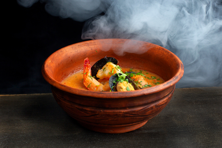 Spicy french soup with seafood in a clay plate. Against the backdrop of beautiful white smoke. Side view.の写真素材
