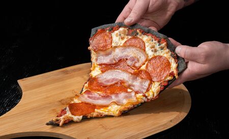 A slice of pepperoni pizza with bacon, on black dough. Lies on a wooden platform.の写真素材