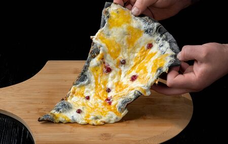 A slice of pizza with cranberries from black dough lying on a wooden board. A man raises a slice of pizza with his hands.の写真素材