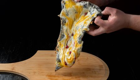 A slice of pizza with cranberries from black dough lying on a wooden board. A man raises a slice of pizza with his hands.の写真素材