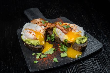 Poached eggs with bacon on toasts. Black bread toast with sesame seeds. Beautiful yolk flows out.の写真素材