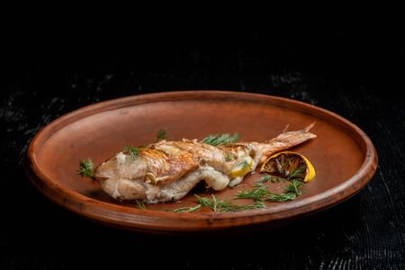 Grilled sea bass on a beautiful clay plate. Sprinkled with dill and garnished with lemon. Side view.