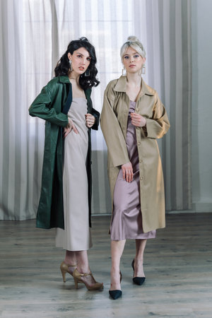 Young girls stands in the studio on the background of curtains. Fashionable modern raincoats are worn over the dresses. Beautiful healthy teeth are visible.の写真素材