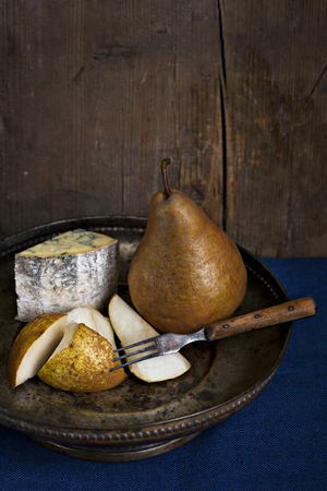 Classic low key still life with Blue cheese with pears on an old fashioned plate against deep blue table cloth and wooden wall. One pear is chopped in pieces and old fashioned fork pricks one piece.の写真素材
