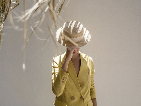 Stylish female in nice jacket hiding face behind straw hat while standing on beige background.