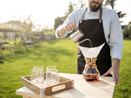 Crop shot of professional man in apron standing at table outdoors in sunlight and pouring water in  coffee maker with filter.の写真素材