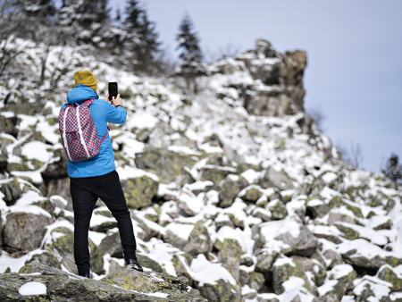 Back view active man in hat and ski jacket taking photo on mobile phone for insta and mounting on stone hill.の写真素材