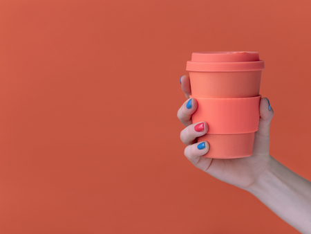 Hand of anonymous female with bright manicure holding takeaway cup of fresh coffee against coral...