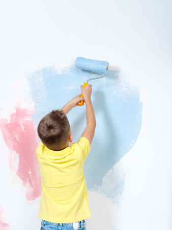 Unrecognizable little cute boy wearing a bright yellow shirt and jeans dyeing the huge white canvas. He paints with blue and rose red paint holding a roller in both hands. New accommodation, renovation or reconstruction concept.の写真素材