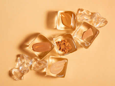 Diagonal composition of samples of liquid foundation, concealer, shimmer, compact powder on artificial ice cubes on beige background. Skin make up products. Summer face makeup, cooling cosmetics concept.の写真素材