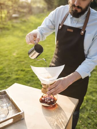 Bearded man pours hot water inside filter coffee utensil. Barista preparing drip coffee outdoor at summertime. Coffee catering. Alternative coffee brewing.の写真素材