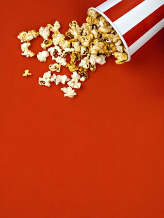 Spilled popcorn in a striped red and white cardboard box on a red background. Cinema, movies and entertainment concept. Top view, copy space.の写真素材