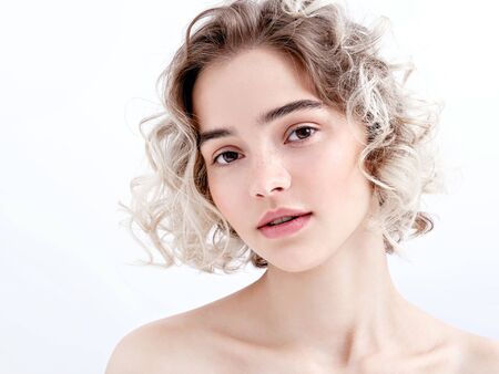 Portrait of a cute young girl with bare shoulders, clean fresh skin and cute curls. Charming model in a soft daylight on white background. Natural feminine beauty. Natural make-up.の写真素材