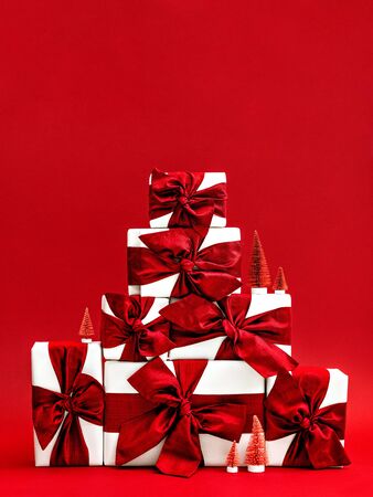Bright red Christmas background. Gift boxes wrapped in white craft paper decorated with handmade ribbons and bows. Pink and red artificial fir-trees beside. Greeting card design. Copy space for text.の写真素材
