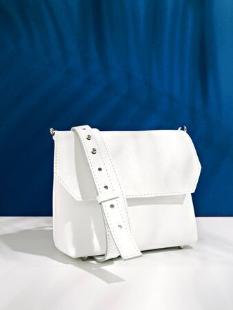 Minimalist stylish female accessory. Trendy white crossbody handbag on deep classic blue background covered with tropic leaves shadow. Color of the year 2020. Summer sale, style, fashion concept.の写真素材
