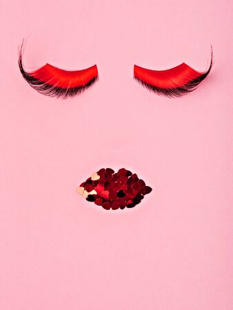 Abstract female face evening or stage makeup. Enormous false red and black lashes, glitter red lips on the pink background. Makeup concept flat lay.の写真素材