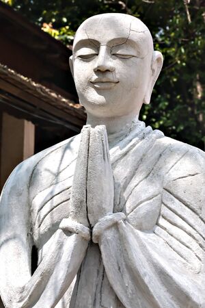 The stone statue of praying Buddhist monk. Paying a respect, praying, relax and deep meditation concept. Buddhist statue with peaceful face. Buddhism symbol.の写真素材