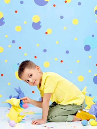 Cheerful little boy plays nose to nose with big pastel coloured origami bunnies in the studio with...