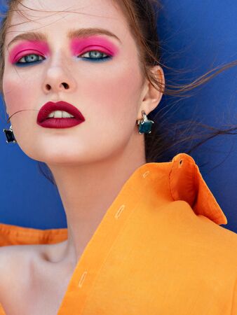 Fashion portrait of stunning woman with bright makeup  and fluttering hair wearing orange shirt. Sensual portrait of red-haired girl on blue background. Summer mood.の写真素材