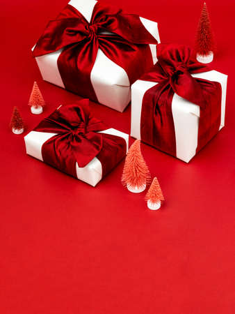Gift boxes wrapped in white paper and tied with luxury wine red ribbon and red Christmas trees on deep red background. Christmas text background for greeting card. Concept of a New year or Christmas.の写真素材