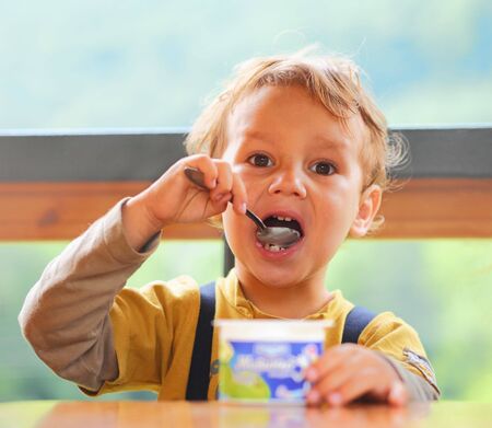 Little boy is eating yogurt, holding a spoon in his hand, sitting at the table .の写真素材