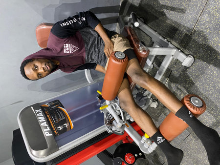 A young Black man is focused on training his legs using a leg extension machine in a commercial gym...
