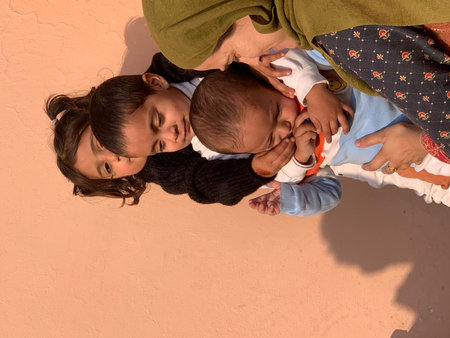 Mother cradles two children against a soft peach wall. Her young daughter rests her head close, eyes half-closed. The baby sleeps soundly, wrapped snugly in light blueの写真素材