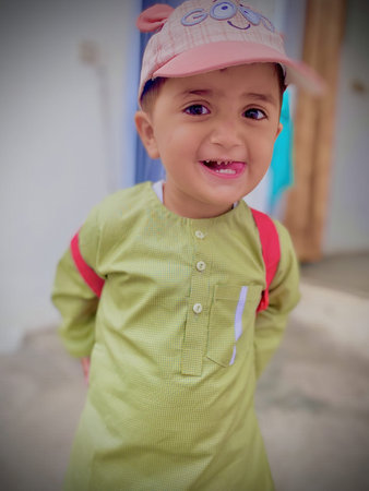 A young boy wearing a green shirt and a pink hat is smiling. He has a backpack on his back