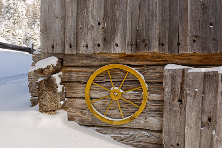 Old yellow wagon wheel hanging on wall to decorate rustic wooden barn wall with cold snow winter scene in Austria, Europeの写真素材