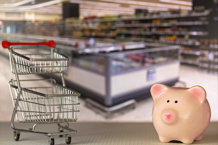 Pink piggy bank pig standing near mini shopping push cart with blurred supermarket backgroundの写真素材