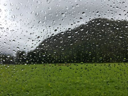 Blurred view of cows on green field, big mountain in rainy day with rain drops splashing windscreenの写真素材