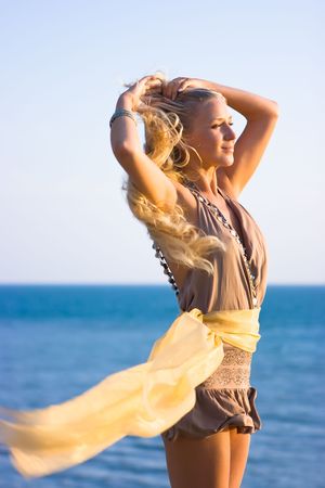 Beautiful blonde girl with transparent gold shawl on the rock and sea in backgroundの写真素材