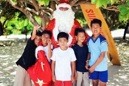 Santa Claus on the beach under a palm tree wishes merry Christmas and gives gifts to cheerful and happy Asian children. Merry Christmas in Polynesiaのeditorial素材