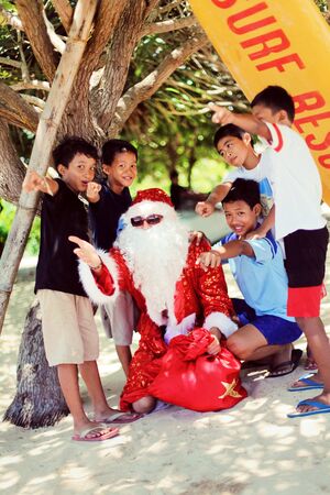 Santa Claus on the beach under a palm tree wishes merry Christmas and gives gifts to cheerful and happy Asian children. Merry Christmas in Polynesiaのeditorial素材