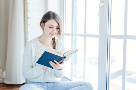 Young woman at home sitting on modern chair in front of window relaxing in her living room reading book and drinking coffee or teaの写真素材