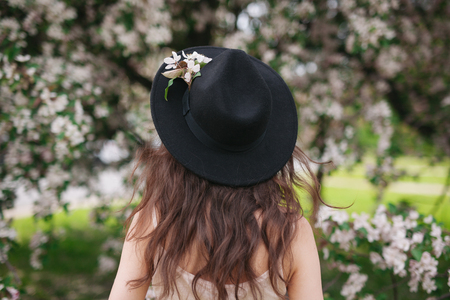 Portrait of beautiful and charming back view of young pretty brunette woman with hands on a hat, on flower field background. Amazing background.の写真素材