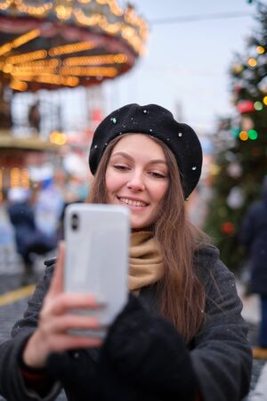 Beautiful young woman taking a selfie with christmas tree behind her with mobile phone camera - Pretty girl enjoying christmas holidays taking a self photography in the city with her smartphoneの写真素材