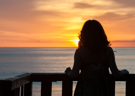 Girl seen from behind standing on a balcony in front of the sea and looking the sunriseの写真素材