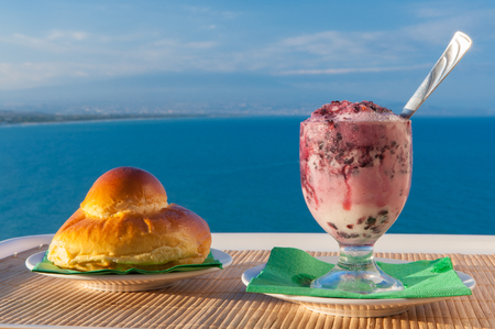 Glass of sicilian granita and a typical warm brioche with sea and mount etna in the backgroundの写真素材