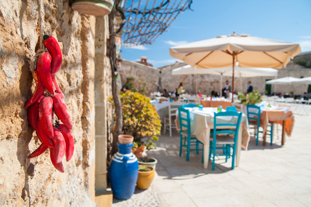 Small pile of fake red peppers hanging on the wall of a typical stone house in the fishing village Marzamemi, Sicily, and the main square with tables of a restaurantの写真素材