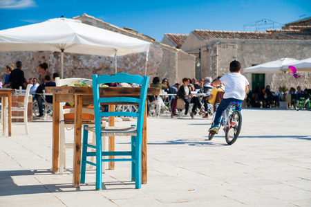 Perspective of the main square of Marzamemi, Sicily, with a wooden table and chairs in the foreground and peolple sitting in a bar in the foregroundの写真素材