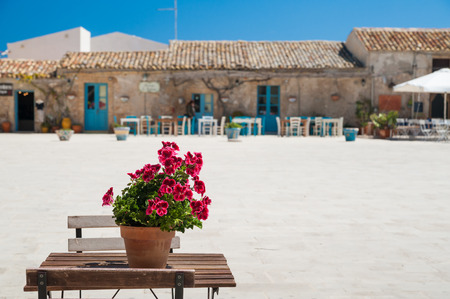 View of the main square in Marzamemi, a picturesque sicilian fishing village, with a flowered vase of geraniums on a small table in the foregroundの写真素材