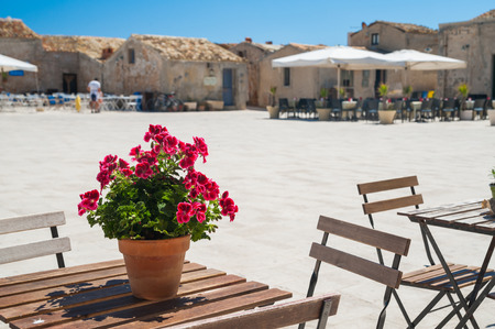 View of the main square in Marzamemi, a picturesque sicilian fishing village, with a flowered vase of geraniums on a small table in the foregroundの写真素材