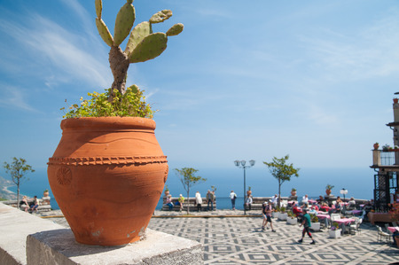 A terracotta vase with a prickly pear and the panoramic square of the small town Castelmola, Sicily, with tourists relaxingの写真素材