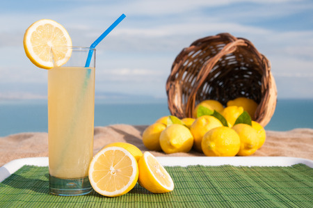 Glass of fresh lemon juice on a piece of Juta with a full wicker basket, blue sea in the backgroundの写真素材