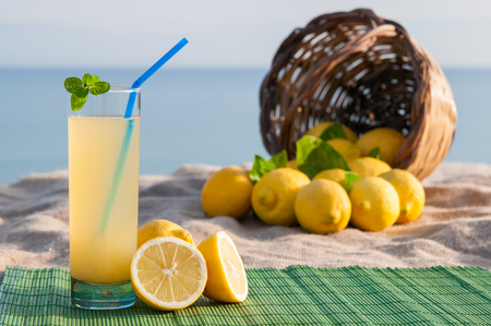 Glass of fresh lemon juice on a piece of Juta with a full wicker basket, blue sea in the backgroundの写真素材