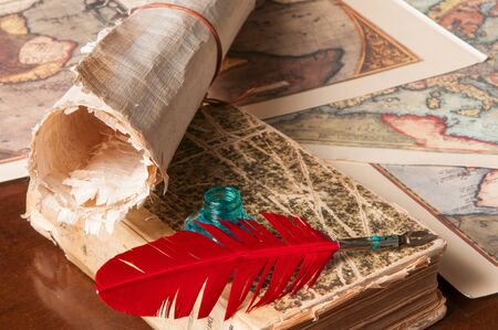 Red quill pen, a green inkwell and a rolled papyrus sheet with some old maps on a wooden tableの写真素材