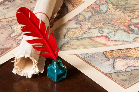 Red quill pen, a green inkwell and a rolled papyrus sheet with some old maps on a wooden table